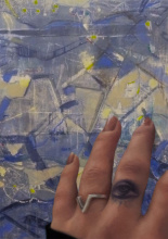 Blau- mattgelbe, abstrakte Malerei mit rechteckigen grafischen Konturen; eine reale Hand ( Foto) auf dem Bild mit einem tätowierten Auge am Mittelfinger sowie einen Silberring mit einem V am Ringfinger , rosa lackierte, fein manekürte Fingernägel